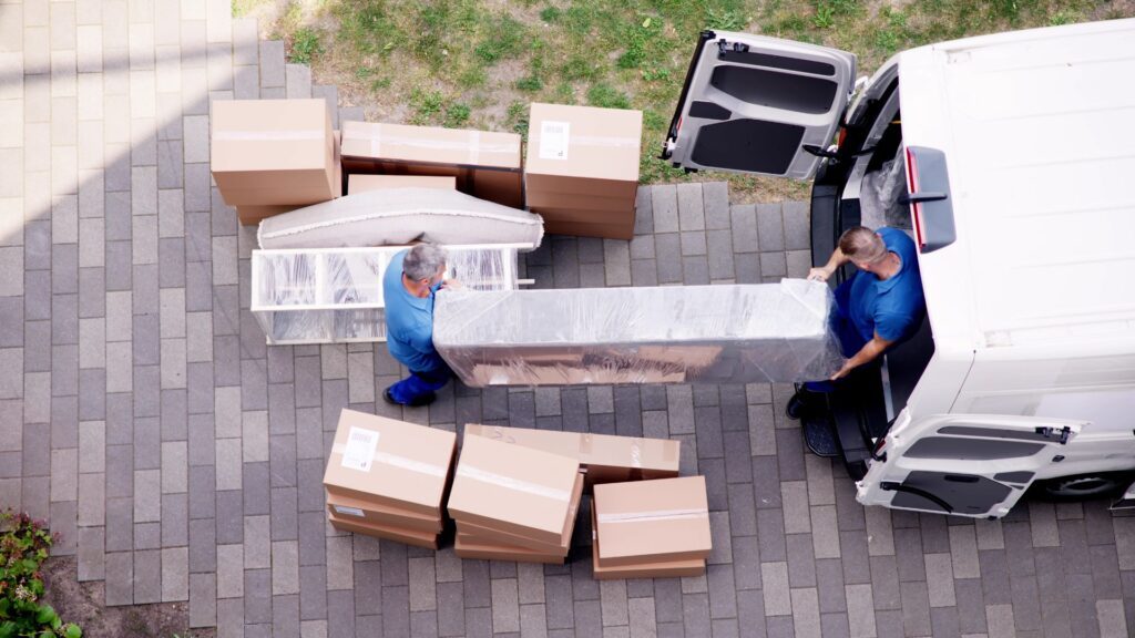 Deux déménageurs en bleu transportant un matelas emballé devant des cartons de débarras et des véhicules de livraison sur un
